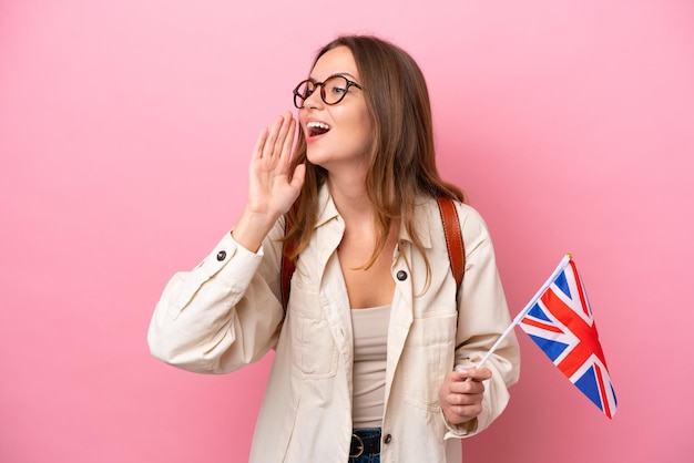 ピンクの背景に分離された英国の旗を保持している若い学生の白人女性が口を横に大きく開いて叫んでいます