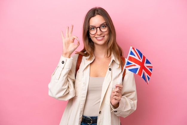 손가락으로 확인 표시를 보여주는 분홍색 배경에 고립 된 영국 국기를 들고 젊은 학생 백인 여자
