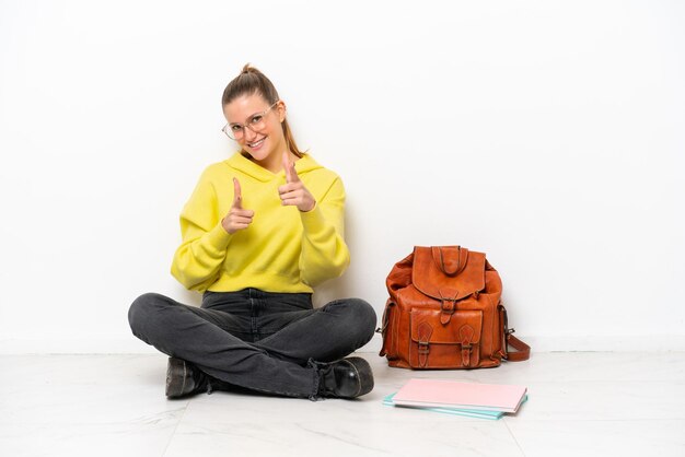 Young student caucasian woman sitting one the floor isolated on white background pointing to the front and smiling