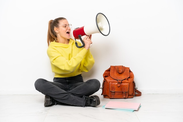 Foto giovane studente donna caucasica seduta uno il pavimento isolato su sfondo bianco gridando attraverso un megafono