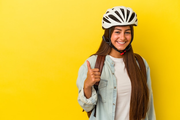 Молодой студент кавказской женщина в велосипедном шлеме, изолированные на желтом фоне, улыбается и поднимает большой палец вверх