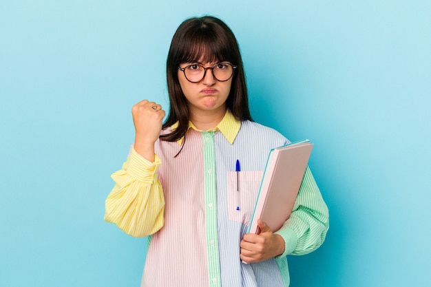 Young student curvy woman holding books isolated on blue background showing fist to camera, aggressive facial expression.