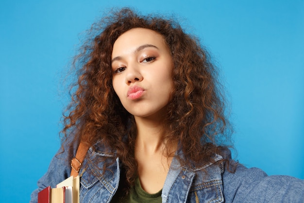 Young student in denim clothes and backpack holds books, sends kiss and makes selfie photo isolated on blue wall