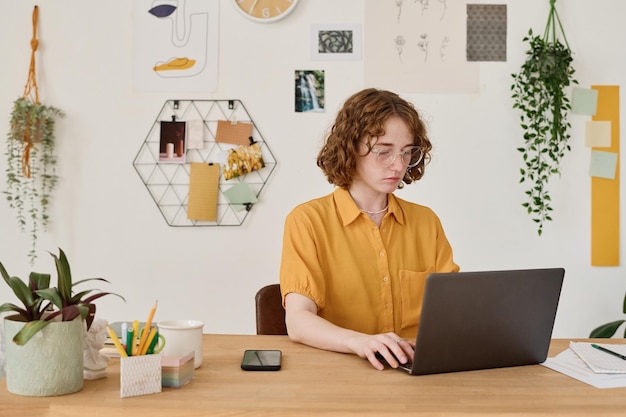 Young student or freelancer in casualwear sitting in front of laptop by desk