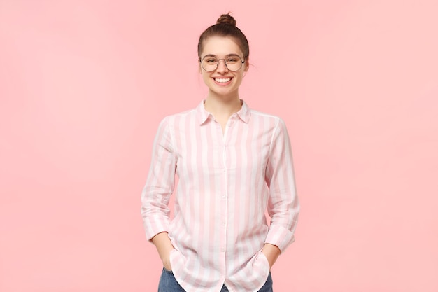 Young student girl dressed in shirt and jeans wearing round glasses and hair tied in bun smiling happily holding hands in pockets