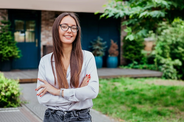 A young student girl in glasses smiles with folded arms against the background of the building