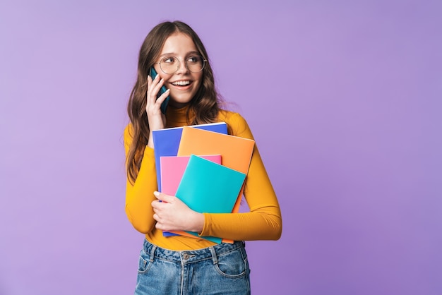 young student girl wearing eyeglasses holding exercise books and talking on cellphone isolated