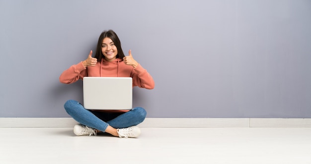 Young student girl with a laptop on the floor giving a thumbs up gesture