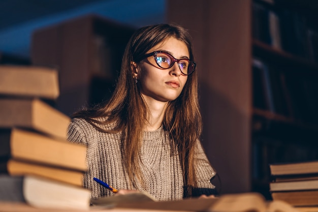 Young student in glasses preparing for the exam. Girl in the evening sits at a table in the library with a pile of books