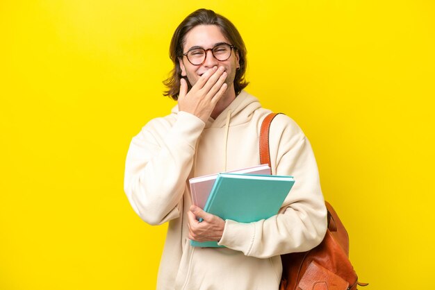 黄色の背景幸せと手で口を覆っている笑顔で分離された若い学生ハンサムな男