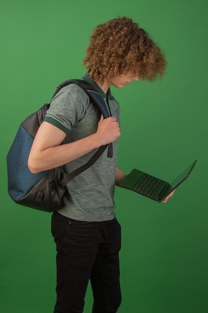 Молодой студент в джинсовой одежде с рюкзаком держит в руках ноутбук Изолированный на зеленом фоне Учеба в университете онлайн Видеосвязь Карантин