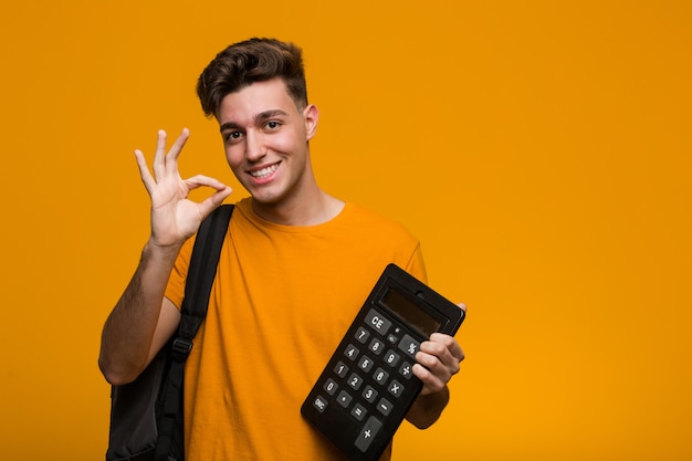 Молодой студент человек, держащий калькулятор удивлен, указывая на себя, широко улыбаясь.