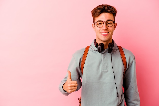 Молодой студент человек изолирован на розовом фоне улыбается и поднимает палец вверх