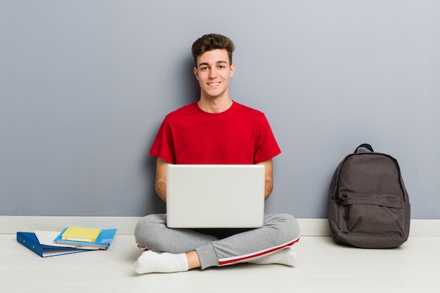 Молодой студент человек сидит на полу своего дома, держа ноутбук