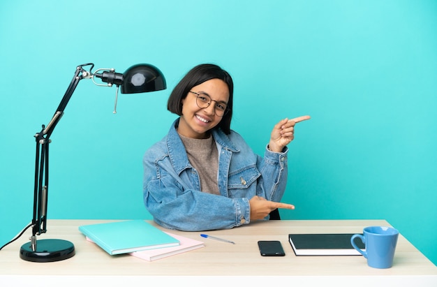 Young student mixed race woman studying on a table pointing finger to the side and presenting a product