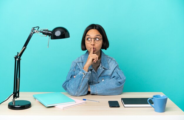 Young student mixed race woman studying on a table showing a sign of silence gesture putting finger in mouth