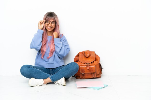 眼鏡と幸せな白い背景で隔離の1つの床に座っているピンクの髪の若い学生混血の女性