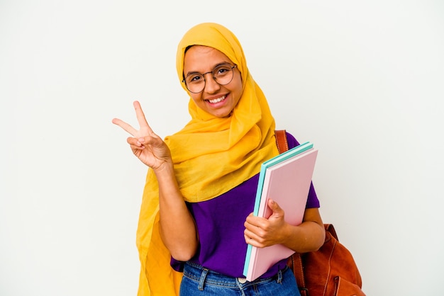 Young student muslim woman wearing a hijab on white joyful and carefree showing a peace symbol with fingers.