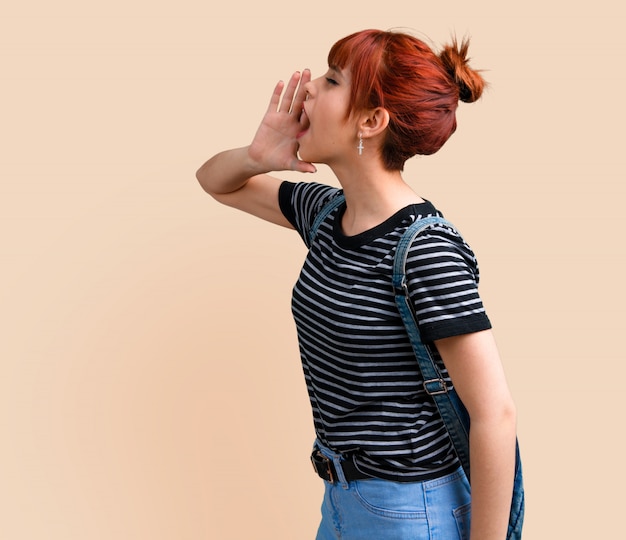 Young student redhead girl shouting with mouth wide open on ocher background.