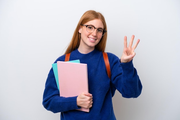 Молодая рыжеволосая студентка на белом фоне счастлива и считает три пальцами