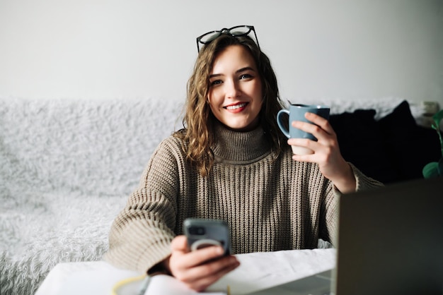 Young student taking a break from online studies enjoying coffee and phone laughing at memes and