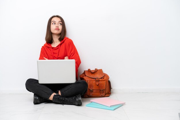 Young student Ukrainian woman sitting one the floor isolated on white background making doubts gesture while lifting the shoulders