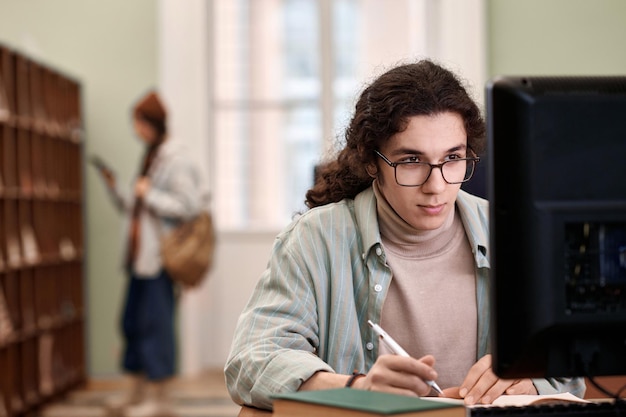 안경을 쓴 젊은 학생이 도서관에서 컴퓨터를 보고 있다