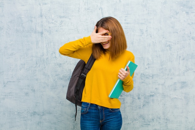 Young student woman covering face with hands peeking between fingers with surprised expression and looking to the side