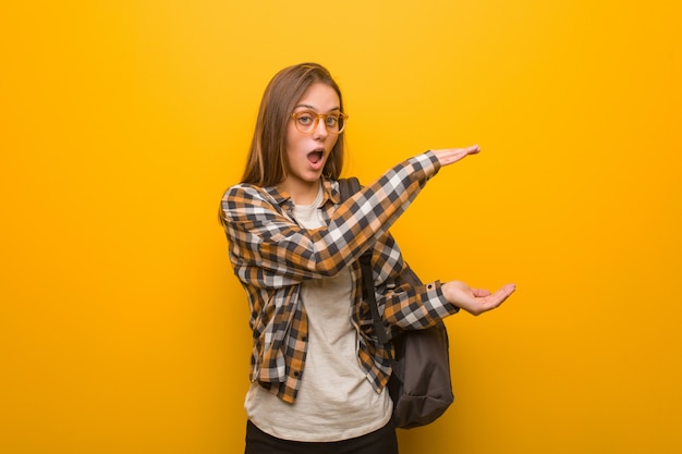 Young student woman holding something very surprised and shocked