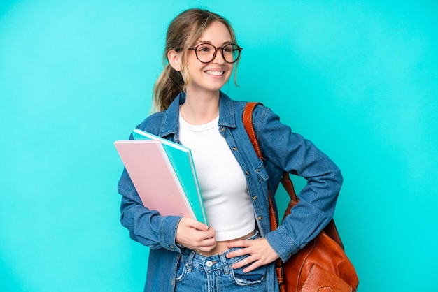 青の背景に腕を腰に当て、笑顔でポーズをとる若い学生の女性