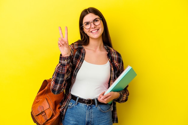 Donna giovane studente isolata sulla parete gialla gioiosa e spensierata che mostra un simbolo di pace con le dita.