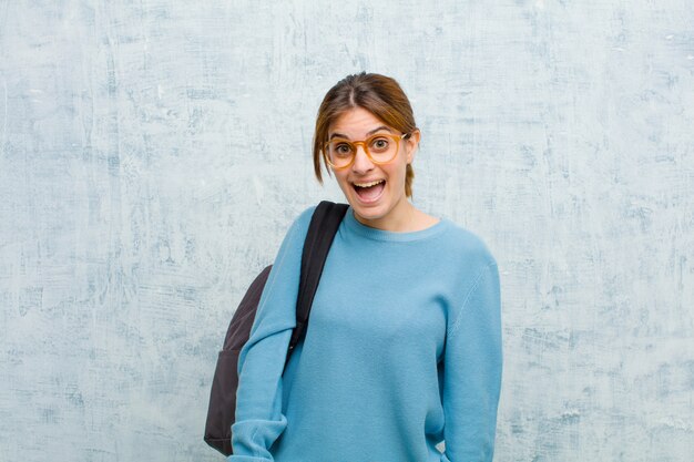 Young student woman looking happy and pleasantly surprised, excited with a fascinated and shocked expression against grunge wall background