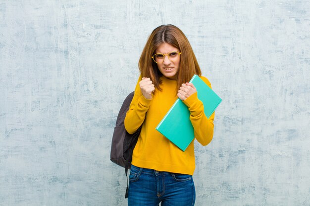 Young student woman shouting aggressively with annoyed, frustrated, angry look and tight fists, feeling furious  grunge wall 