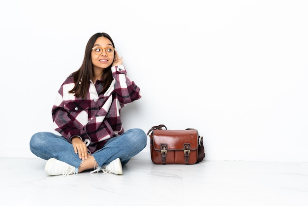 Young student woman sitting on the floor listening to something by putting hand on the ear