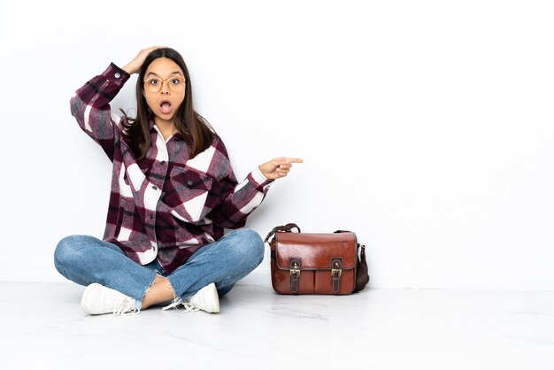 Young student woman sitting on the floor surprised and pointing finger to the side