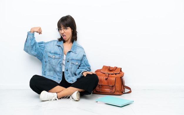 Young student woman sitting one the floor isolated on white background doing strong gesture