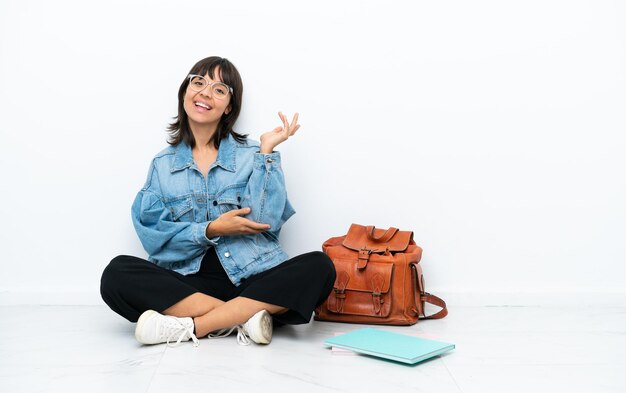 白い背景で隔離された床の1つに座っている若い学生の女性は、来て招待するために手を横に伸ばします