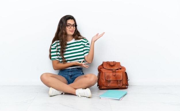 Young student woman sitting one the floor isolated on white background extending hands to the side for inviting to come