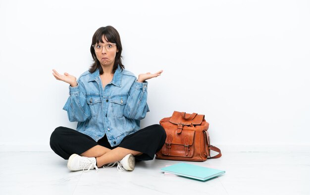 Young student woman sitting one the floor isolated on white background having doubts while raising hands