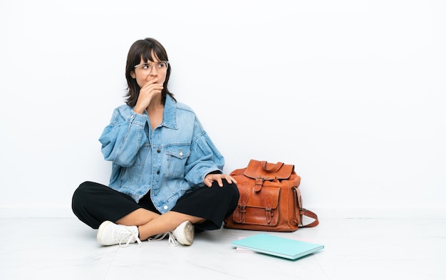 Young student woman sitting one the floor isolated on white background having doubts and with confuse face expression