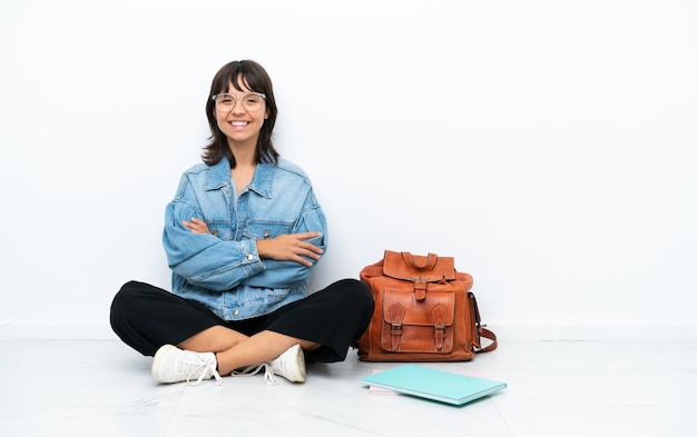 Young student woman sitting one the floor isolated on white background keeping the arms crossed in frontal position