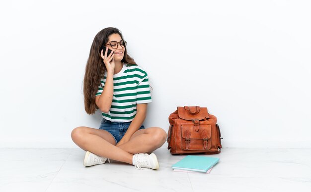 Young student woman sitting one the floor isolated on white background keeping a conversation with the mobile phone