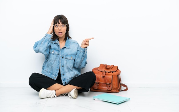 白い背景で隔離された床の1つに座っている若い学生の女性は驚いて、人差し指を横に向けています