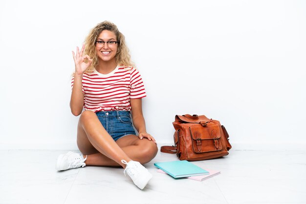 Young student woman sitting one the floor showing ok sign with fingers