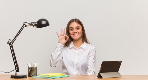 Young student woman working on her desk cheerful and confident doing ok gesture