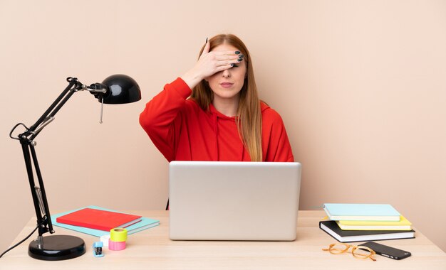 Young student woman in a workplace with a laptop covering eyes by hands