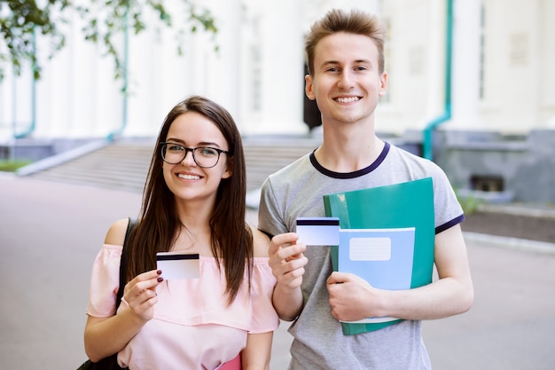 Young students near university with books, exercise books holding credit cards and smiling to camera