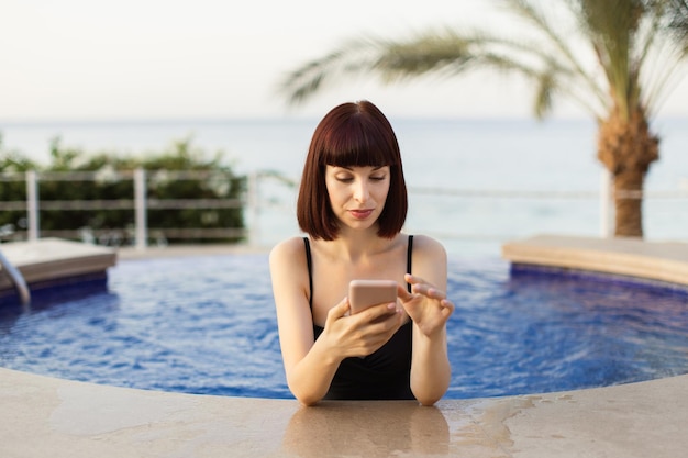 Young stunning woman browsing internet on smartphone at outdoor tropical swimming pool