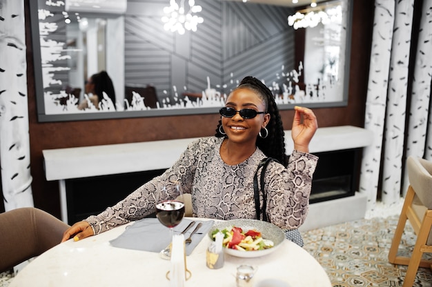 若いスタイリッシュなアフリカ系アメリカ人の女性は、黒いサングラスをかけ、レストランに座って、ワインと一緒に健康的な食事を楽しんでいます。