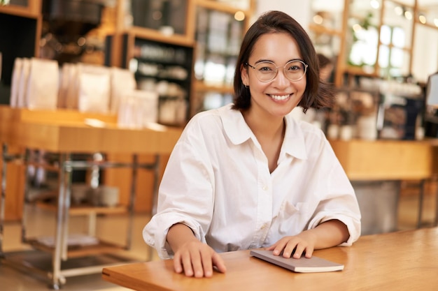 カメラに微笑んでノートを持ってカフェに座っている眼鏡をかけた若いスタイリッシュなアジア人女性の経営者